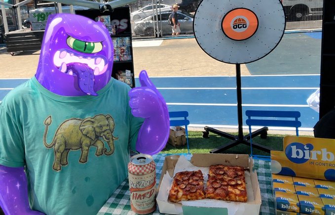 An inflatable purple monster mascot gives a thumbs up while wearing an elephant-printed shirt, sitting at a tailgate table featuring a pizza box, a "Happy" beer can, Birra Moretti cardboard signage, and an OCG-branded target, humorously capturing the celebratory "OGs flipping" victory pose.