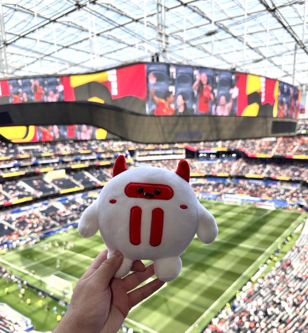Hand holding a white plush mascot with red horns and features, photographed inside a large stadium with a packed crowd and a massive screen displaying performers.