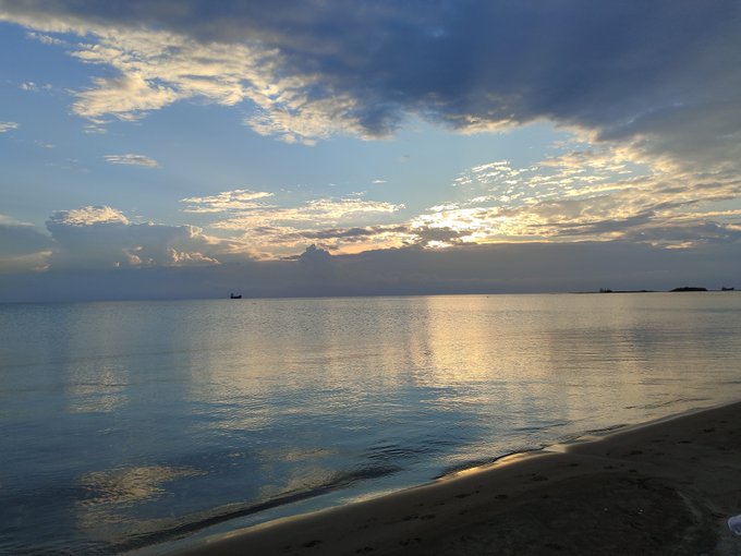 A serene beach scene at sunset or sunrise, with dramatic clouds, golden sunlight reflecting on calm ocean waters, and a small boat visible on the horizon.