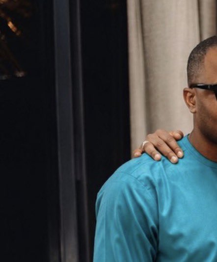 A man in a teal sweater and sunglasses rests his hand on someone's shoulder, partially framed against a dark background and cream curtains. The intimate gesture and partial composition create a sense of connection, leaving the full scene to the viewer's imagination.
