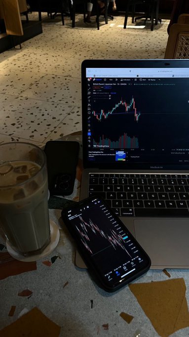A laptop and smartphone display financial candlestick charts, with “British Pound / Japanese Yen (GBP/JPY)” visible on the laptop. A glass of iced drink, a phone, and cables rest on a café table.