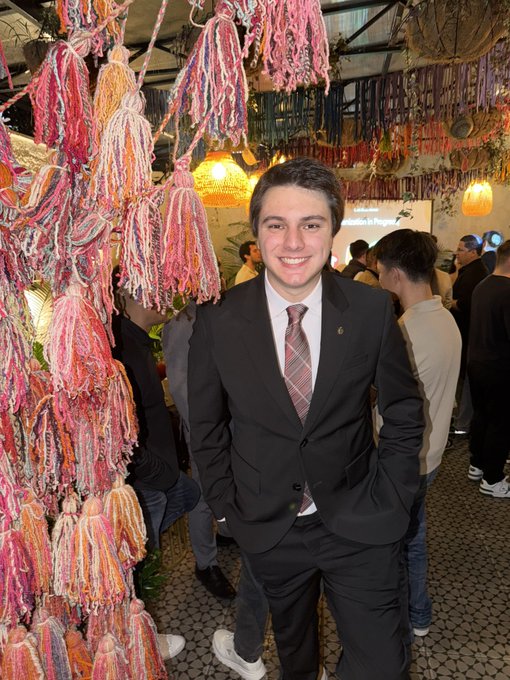 A smiling young man in a dark suit with a plaid tie posing at what appears to be an event, standing next to colorful pink and orange yarn tassel decorations.