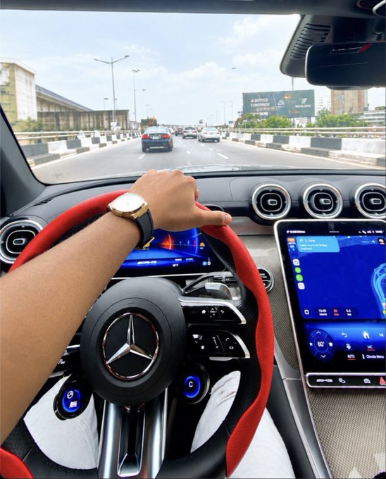 First-person view from the driver's seat of a Mercedes-Benz, showing a hand on a red-accented steering wheel and dual infotainment screens displaying navigation.