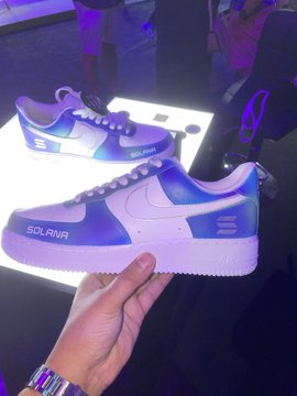 A hand holding up a pair of white and purple sneakers branded 'SOLANA' displayed against an illuminated background.