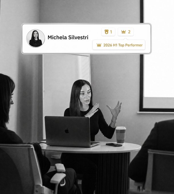 A black-and-white image shows a woman with long dark hair gesturing while seated at a round table with a laptop (Apple logo visible), engaged in conversation with colleagues. An overlay displays "Michela Silvestri" with badges reading "1", "2", and "2026 H1 Top Performer." The caption highlights her recognition as a Top Performer at Binance for 2026, celebrated briefly before returning to her daily work of managing Telegram messages and automation.
