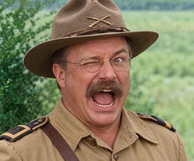 A man with a thick mustache, round spectacles, and a wide-brimmed campaign hat with crossed sabers is shouting with an exaggerated, almost manic expression. The image evokes Theodore Roosevelt's iconic Rough Rider persona, visually reinforcing the "Roughrider Coin" reference.