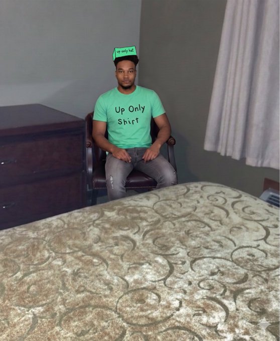 A young man wearing a green t-shirt with the text 'Up Only Shirt' and a matching 'Up Only hat' sits in a chair in a hotel room with a large bed in the foreground.