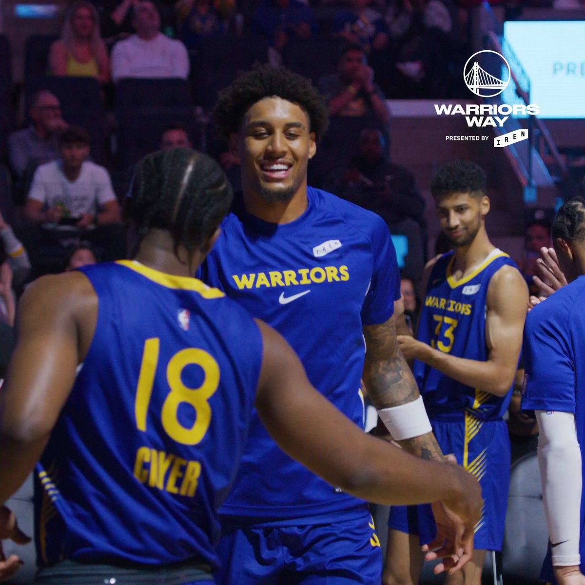 Golden State Warriors players, including #18 and #73, smile and greet each other in their blue and yellow jerseys during a game, with the "WARRIORS WAY presented by IREN" logo visible in the corner, promoting the new season of the docuseries covering the team's summer journey from golf outings and Draft Night to their Summer League championship.