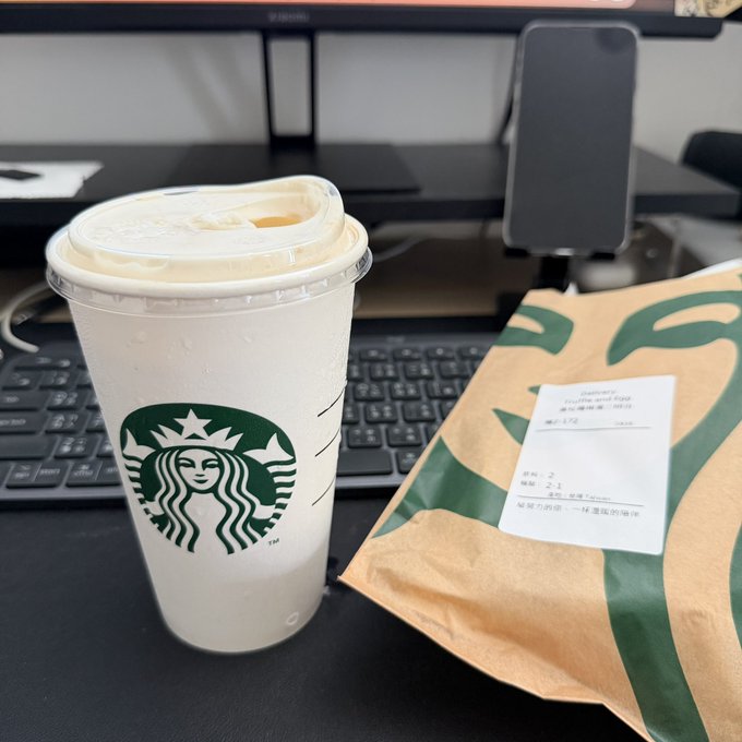 A Starbucks iced drink in a clear plastic cup sits next to a brown paper bag with a receipt reading "mushie sandw" and other order details, placed on a desk with a keyboard and monitor. The image captures the classic fall "first cup of the season" moment referenced in the caption, with the iced beverage representing the autumn Starbucks treat.