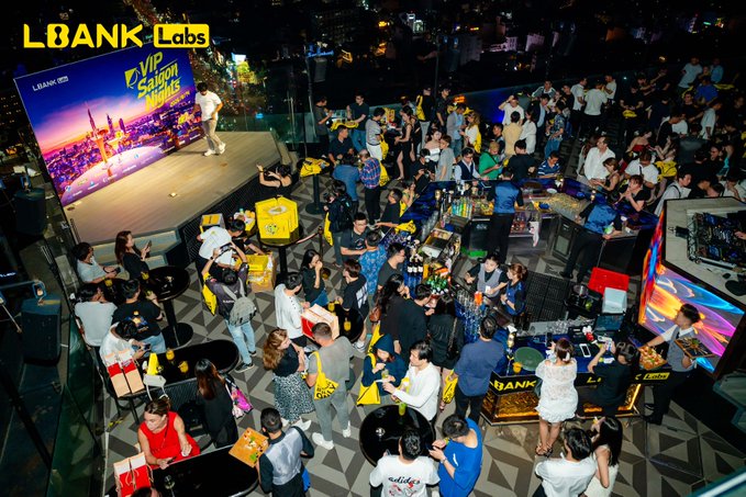 An overhead view of a crowded rooftop event hosted by LBank Labs, featuring attendees, an 'E-VIP Saigon Summer' stage display, and yellow branded merchandise bags.