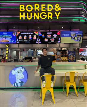 A young man sits on a stool at a brightly lit 'Bored & Hungry' food kiosk/counter, which features KAWS-style character artwork and neon signage.