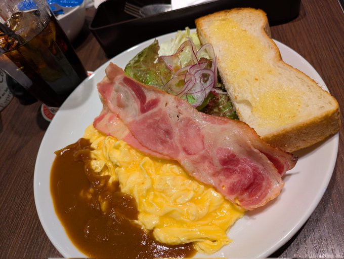 A breakfast plate featuring scrambled eggs with demi-glace sauce, crispy bacon, salad with red onion, toasted bread, and a glass of iced coffee on a wooden table.