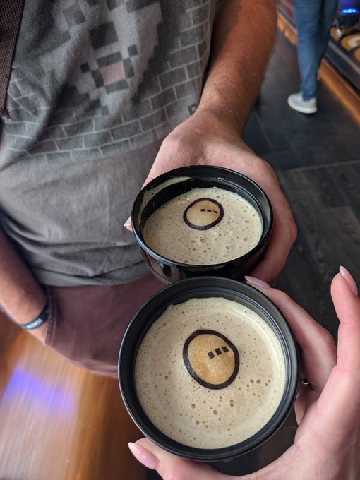Two people clink black cups of a latte-like beverage together, with coffee art on the foam surface.