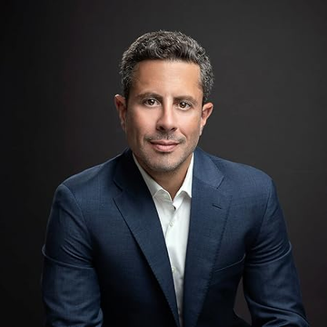 A professional headshot of a middle-aged man in a dark suit jacket and white shirt against a dark gray background.