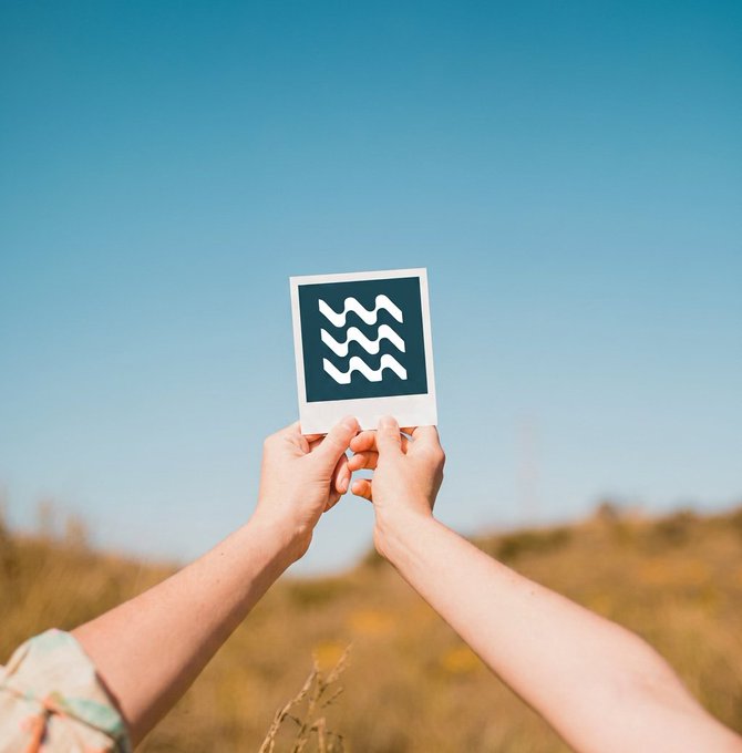 Two hands holding up a Polaroid-style photo with an Aquarius zodiac symbol against a backdrop of a blue sky and grassy field.