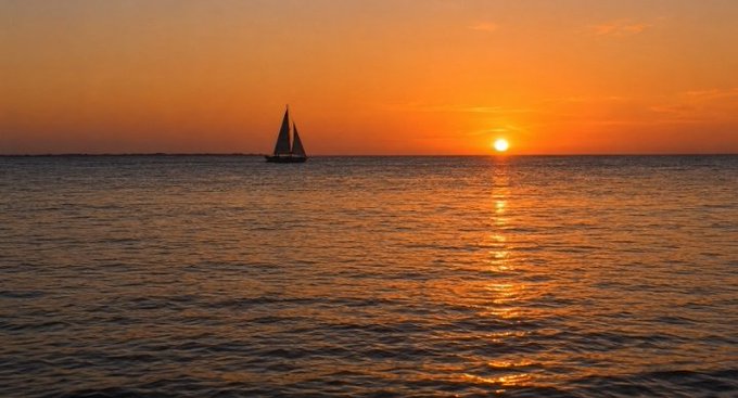 A serene sunset over the ocean with a sailboat silhouetted against an orange sky, the sun's reflection shimmering on the calm water.