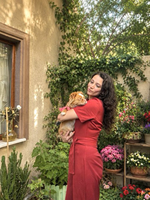 A woman with long curly hair wearing a red dress, smiling while holding a ginger cat in a lush garden setting with flowers and greenery.