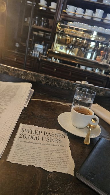 A newspaper clipping with the headline "SWEEP PASSES 20,000 USERS" sits on a marble cafe table next to an espresso cup, saucer, spoon, and a glass of water, with shelves of cups visible in the background.