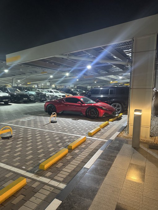 A red Toyota GR Supra sports car is parked at night under a covered parking structure, with yellow parking blocks in the foreground and other vehicles visible in the background.