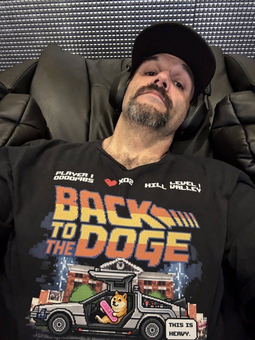 A bearded man wearing a black cap and headphones poses for a selfie while wearing a black t-shirt with a pixel-art design that reads 'BACK TO THE DOGE' with 'PLAYER 1 DOGE1985', 'XOR', and 'LEVEL VALLEY' text, and a caption reading 'THIS IS HEAVY.'