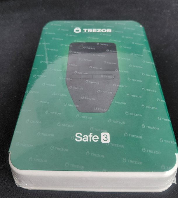 A sealed retail box for a Trezor Safe 3 hardware cryptocurrency wallet, featuring a green design with repeating Trezor logos and an image of the device.