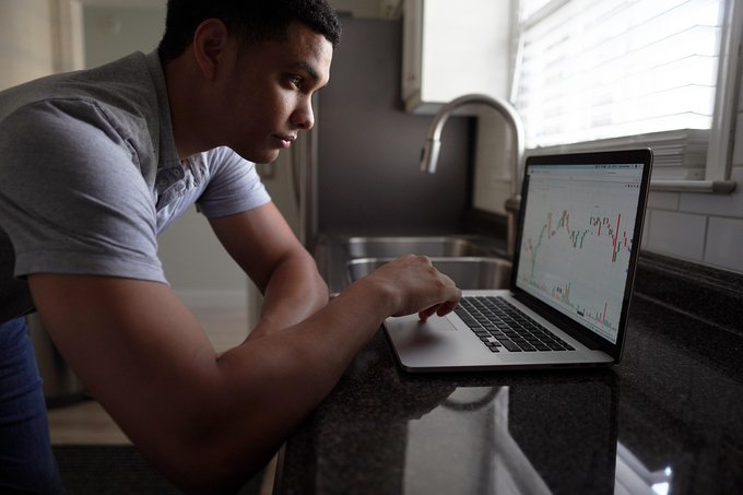 A man in a gray polo shirt leans over a kitchen counter studying a stock chart on his laptop screen.