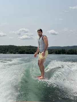 A man in a life vest and yellow shorts wake-surfs barefoot behind a boat, balancing confidently as he glides over the wake with green hills in the distance. The image playfully contrasts his focused, athletic stance with the post's humorous claim of competing against his @doodles toy.