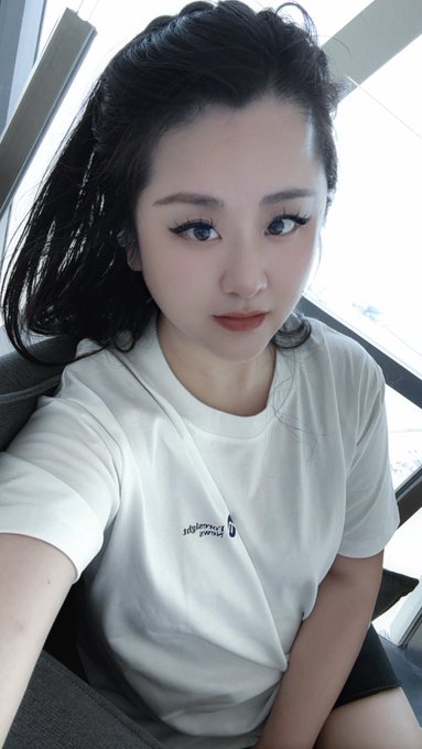 A young woman with long dark hair takes a selfie wearing a white t-shirt, sitting indoors near a window.