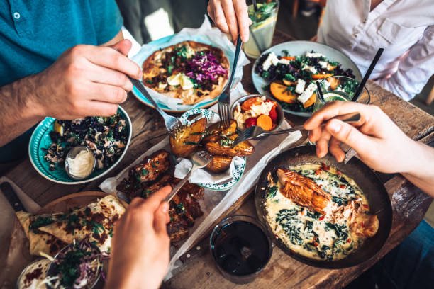 Four people share a wooden table filled with various colorful, vegetable-rich dishes including salads, roasted potatoes, and greens, with hands holding forks and drinks around the meal.