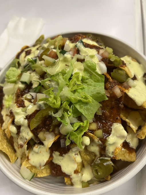 A bowl of loaded nachos topped with lettuce, jalapeños, cheese, and a creamy white sauce drizzle, served on a white countertop.