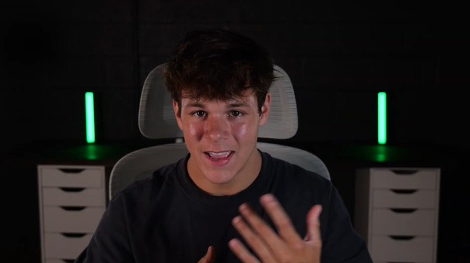 A young man with dark wavy hair, wearing a black t-shirt, speaks directly to the camera while gesturing with his hand, with glowing green light tubes and white drawer units visible in the dark background. The image appears to be a still from his YouTube video, where he discusses his bullish thesis on $ANSEM and predicts that @blknoiz06 will become the "MrBeast of Crypto."