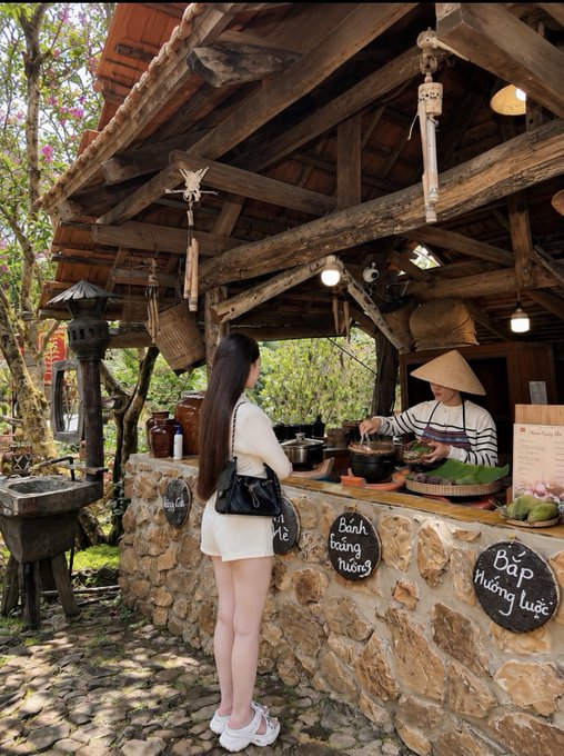 A woman with long dark hair stands at a rustic outdoor kitchen counter while a chef in a conical hat prepares food, with chalkboards displaying Vietnamese menu items like 'Bánh tráng nướng' and 'Bắp nướng'.