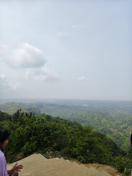 The image shows a person standing at the top of a staircase overlooking a beautiful landscape with mountains, trees, and a clear blue sky. The person appears to be taking a photo or enjoying the view from this elevated position. There are no texts or other objects in the scene that provide information about the post's content.