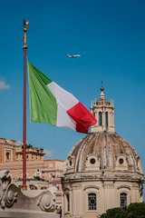 The image shows the Italian flag waving beside a domed building in Rome, symbolizing Italy as UniCredit, the country's second-largest bank, considers entering the crypto custody and brokerage space, marking a step toward institutional adoption of tokenized investments.