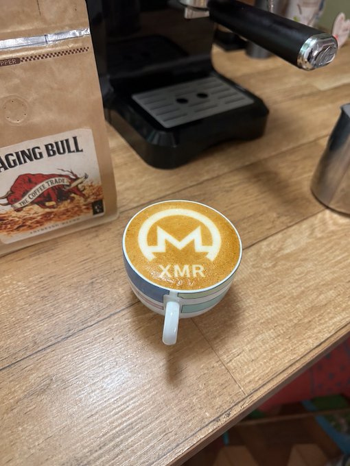 A cup of coffee with latte art featuring the Monero (XMR) cryptocurrency logo, sitting on a wooden table next to a bag of coffee labeled 'RAGING BULL'.