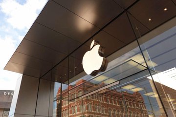The image shows a large glass window of an Apple store, with the iconic Apple logo prominently displayed above it. The store's name is also visible below the logo. In the background, there are several buildings, including one that appears to be an old brick building. The image also contains text that reads "Five of the biggest names in tech just helped the DOJ dismantle a Southeast Asia scam empire in four days."