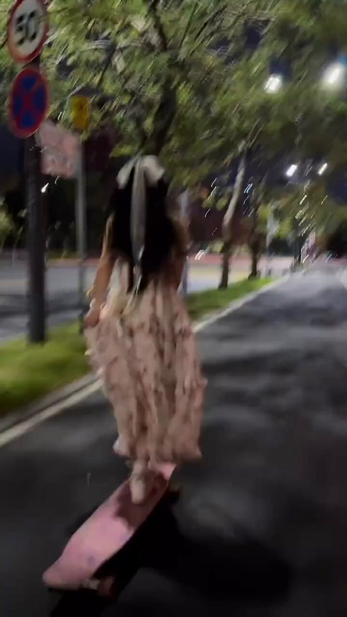 The image shows a woman wearing a white dress riding a pink skateboard down a street at night, with trees lining both sides of the road. The photo has been edited or filtered to emphasize certain elements while omitting others.