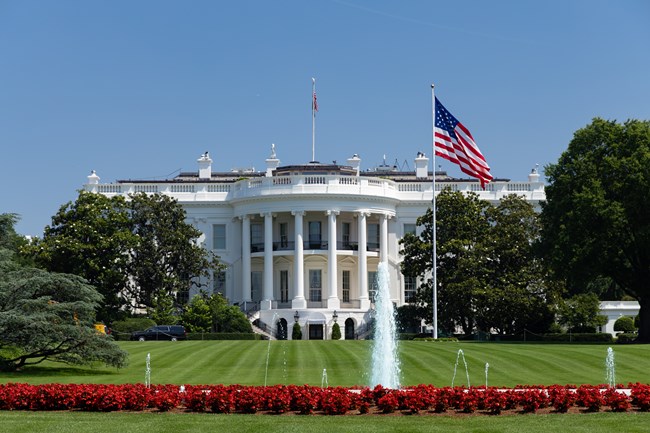 The White House stands prominently with its American flag waving under a clear blue sky, as the administration pushes for the Clarity Act ahead of a crucial Senate vote this September. The image symbolizes the seat of executive power where the crypto summit and legislative efforts are being coordinated.