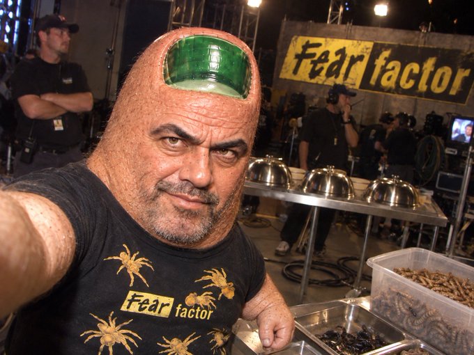 A man in a Fear Factor t-shirt with a green shaved design on his head poses in front of the Fear Factor set.