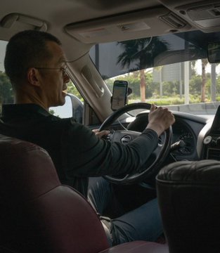 An older man with glasses sits in the driver's seat of a car with a reddish interior, holding a smartphone mounted on the dashboard while his hands are on the steering wheel.
