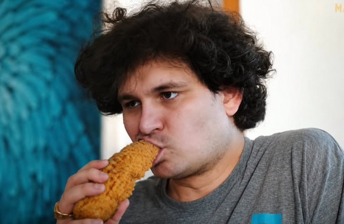 A young man with curly dark hair eats a piece of fried chicken in front of a blue patterned background.