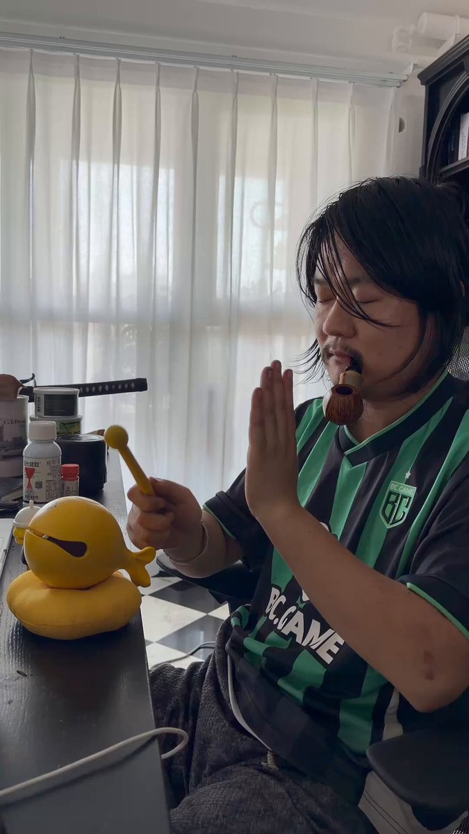 The image features a person sitting at a table with a yellow toy snake and a green toy car. The person appears to be eating food from a plate while holding the toy snake, possibly as part of a playful moment or for entertainment purposes. The background shows a window that allows natural light into the room, creating a cozy atmosphere.
