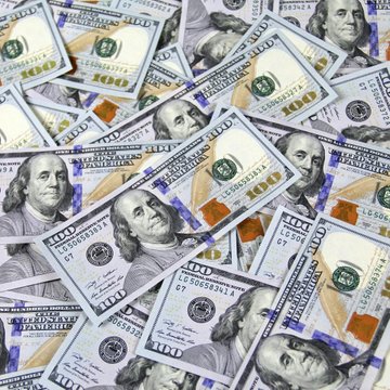 A pile of crisp U.S. $100 bills featuring Benjamin Franklin's portrait is scattered across the frame, visually representing the strengthening dollar highlighted in the post, which cites Glassnode's analysis of the dollar's renewed dominance pressuring Bitcoin and the broader crypto market.