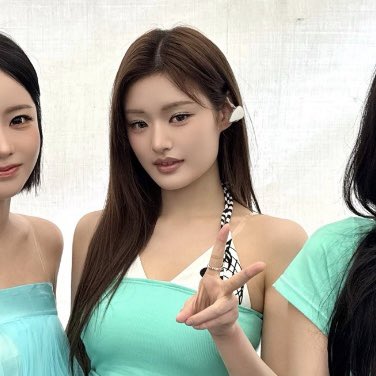 Three young women posing together, with the central figure featuring long brown hair, a hair clip, and a turquoise top making a finger heart gesture.
