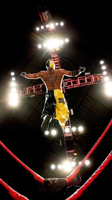 A dramatic action shot of a wrestler wearing yellow trunks and a mask leaping through the air in an arena, illuminated by bright stage lights from above, with the championship belt visible in the background.