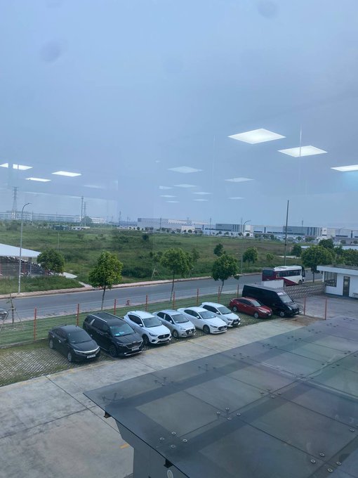 A view from a window overlooking a parking lot with several cars, a road, trees, and industrial buildings under an overcast sky.
