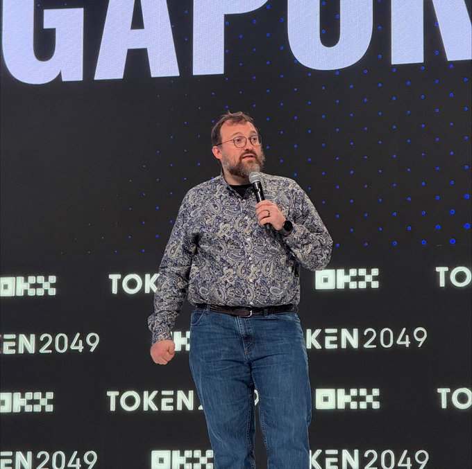 A bearded man with glasses, wearing a patterned shirt and jeans, speaks into a microphone on stage at TOKEN2049, with "TOKEN2049" and "OKX" branding visible on the backdrop behind him. The scene reflects the conference mentioned in the post, where AI agents from Midnight City had human proxies walking the floor on their behalf.