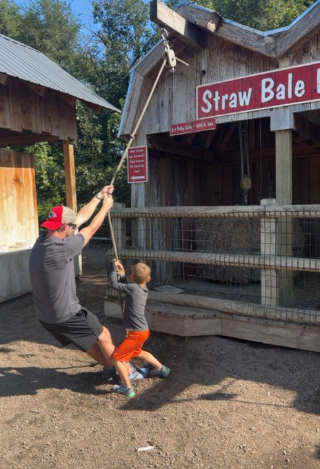 A man and his young son pull on a rope together at a "Straw Bale" attraction, straining to lift the heavy load — just like a crypto investor struggling to recover from underwater altcoin bags, where the burden feels enormous and the payoff remains painfully out of reach.
