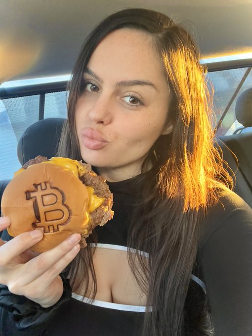 A woman sitting in a car at sunset, posing and pouting her lips while holding a hamburger branded with the Bitcoin (₿) logo.