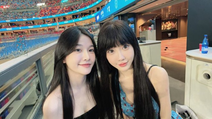 A selfie of two young women with long dark hair smiling at the camera inside a large stadium, with tiered seating and a sports field visible in the background.